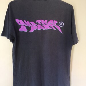 Może przedstawiać: Szary T-shirt z fioletowym nadrukiem, na kt&oacute;rym widnieje napis "Take That & Party" z małym k&oacute;łkiem i krzyżem w środku.