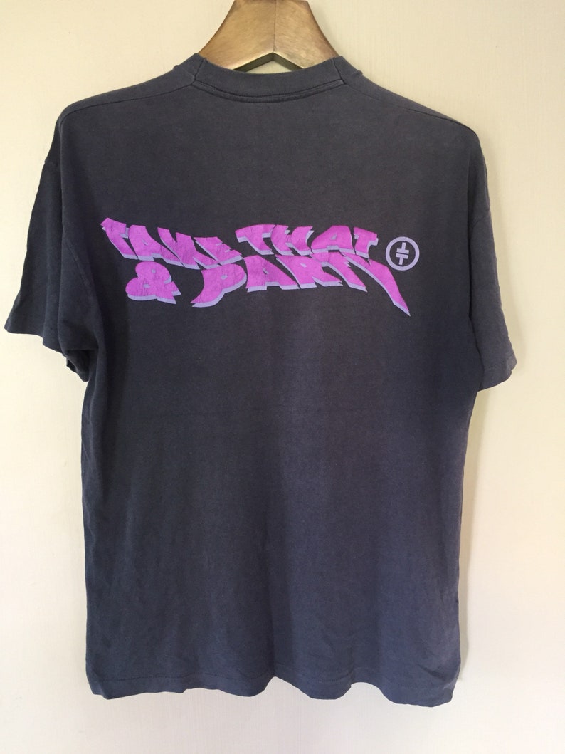 Może przedstawiać: Szary T-shirt z fioletowym nadrukiem, na kt&oacute;rym widnieje napis "Take That & Party" z małym k&oacute;łkiem i krzyżem w środku.