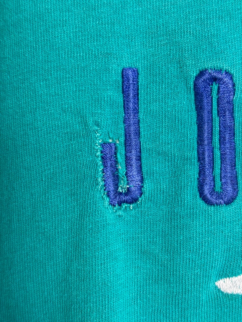 K&ouml;nnte beinhalten: Nahaufnahme eines t&uuml;rkisfarbenen Stoffes mit den blau gestickten Buchstaben "JO". Der Buchstabe "J" weist einige Abnutzungserscheinungen auf. Der Stoff hat ein strukturiertes Aussehen.