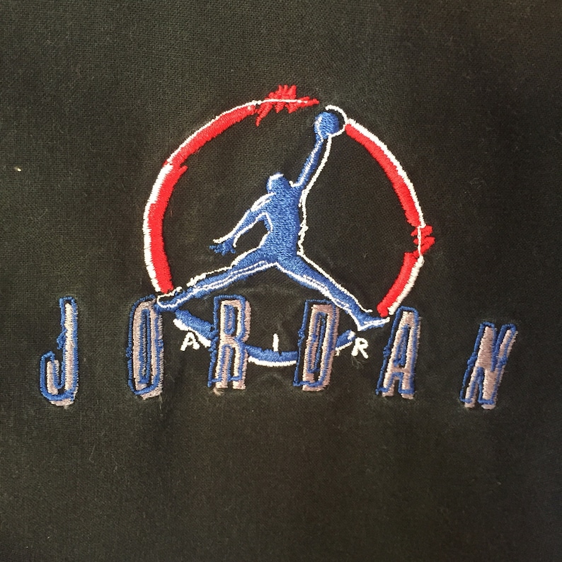 Puede incluir: Sudadera negra con un logotipo bordado en rojo, blanco y azul de un jugador de baloncesto haciendo un mate. El logotipo est&aacute; rodeado de un c&iacute;rculo con la palabra "JORDAN" escrita en letras azules y grises debajo.