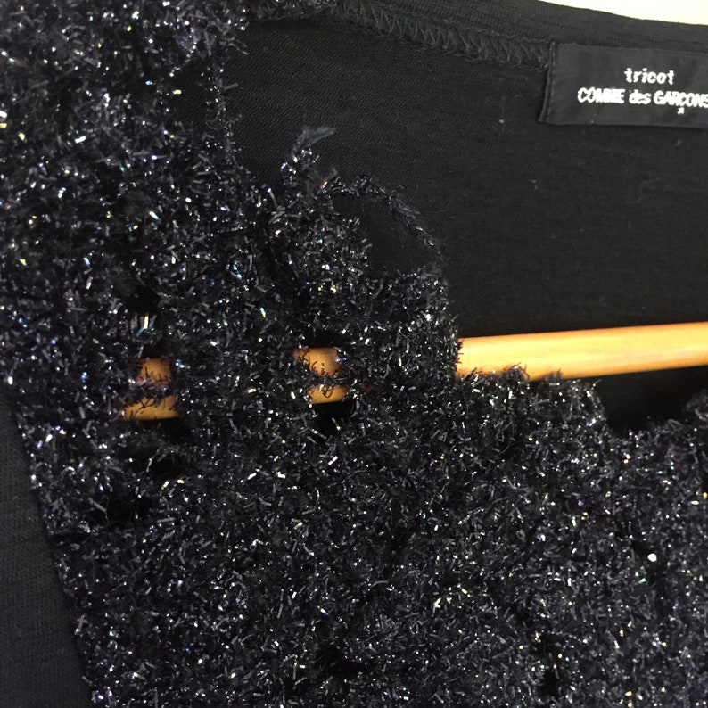 May include: Black shirt with a sparkly, textured trim around the neckline. The shirt has a label that reads "tricot COMME des GARCONS".