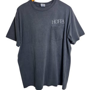 Puede incluir: Una camiseta gris carbón con un bolsillo en el pecho izquierdo. La palabra "HOFFA" está impresa en blanco en la parte superior derecha del pecho. La camiseta está colgada de una percha de madera.