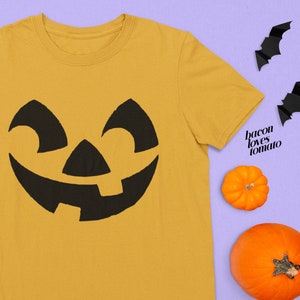Puede incluir: Una camiseta amarilla con un diseño de cara de calabaza negra. La camiseta está rodeada de pequeñas calabazas naranjas y murciélagos negros sobre un fondo morado claro.