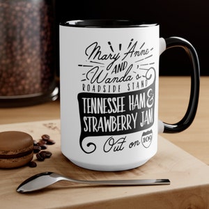 May include: White ceramic mug with a black rim and handle. The mug features black text that reads "Mary Anne & Wanda's Roadside Stand Tennessee Ham & Strawberry Jam Out on 109".
