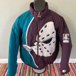 Puede incluir: Una chaqueta vintage Starter de color morado y turquesa con un logotipo blanco de Mighty Ducks y un pato de dibujos animados con una máscara de hockey en la parte delantera. La chaqueta tiene una cremallera y un logotipo de Starters en la manga.