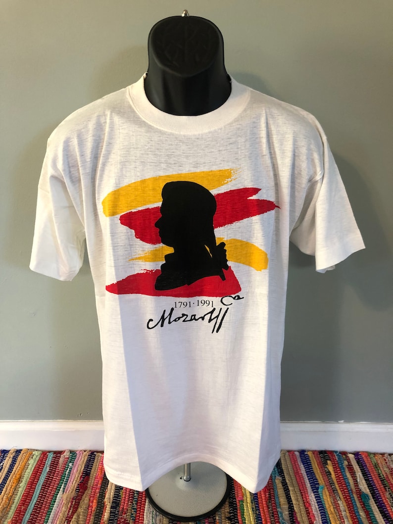 Pu&ograve; includere: Maglietta bianca con una silhouette nera di Wolfgang Amadeus Mozart, pennellate rosse e gialle e il testo "1791-1991" e "Mozart" in nero.