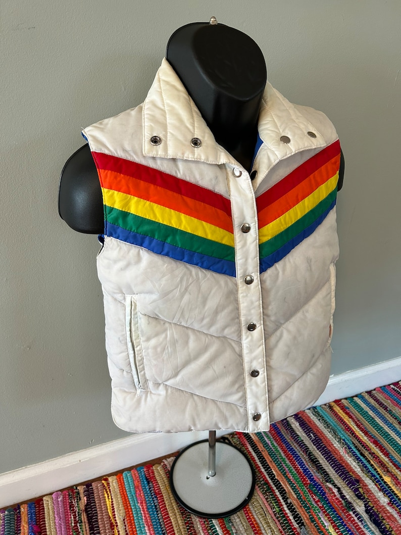 May include: White puffer vest with a rainbow stripe design. The vest has a snap closure and a stand-up collar.