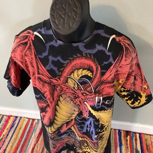 May include: Black t-shirt with a red, yellow, and blue dragon graphic. The dragon is breathing fire and has its wings spread out. The background is a dark blue with clouds.