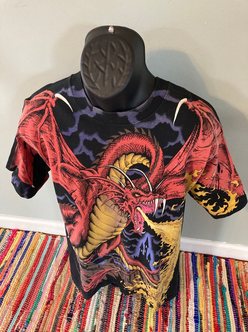 May include: Black t-shirt with a red, yellow, and blue dragon graphic. The dragon is breathing fire and has its wings spread out. The background is a dark blue with clouds.