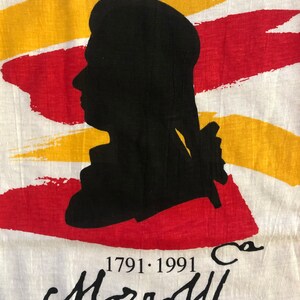 Pu&ograve; includere: Una maglietta bianca con una silhouette nera di una testa e spalle di una persona. La silhouette &egrave; posizionata su uno sfondo di pennellate rosse e gialle. Il testo "1791.1991" e una firma che recita "Mozart" sono stampati sotto la silhouette.