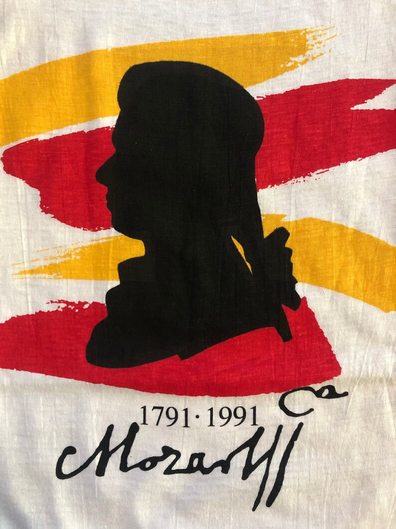 Pu&ograve; includere: Una maglietta bianca con una silhouette nera di una testa e spalle di una persona. La silhouette &egrave; posizionata su uno sfondo di pennellate rosse e gialle. Il testo "1791.1991" e una firma che recita "Mozart" sono stampati sotto la silhouette.