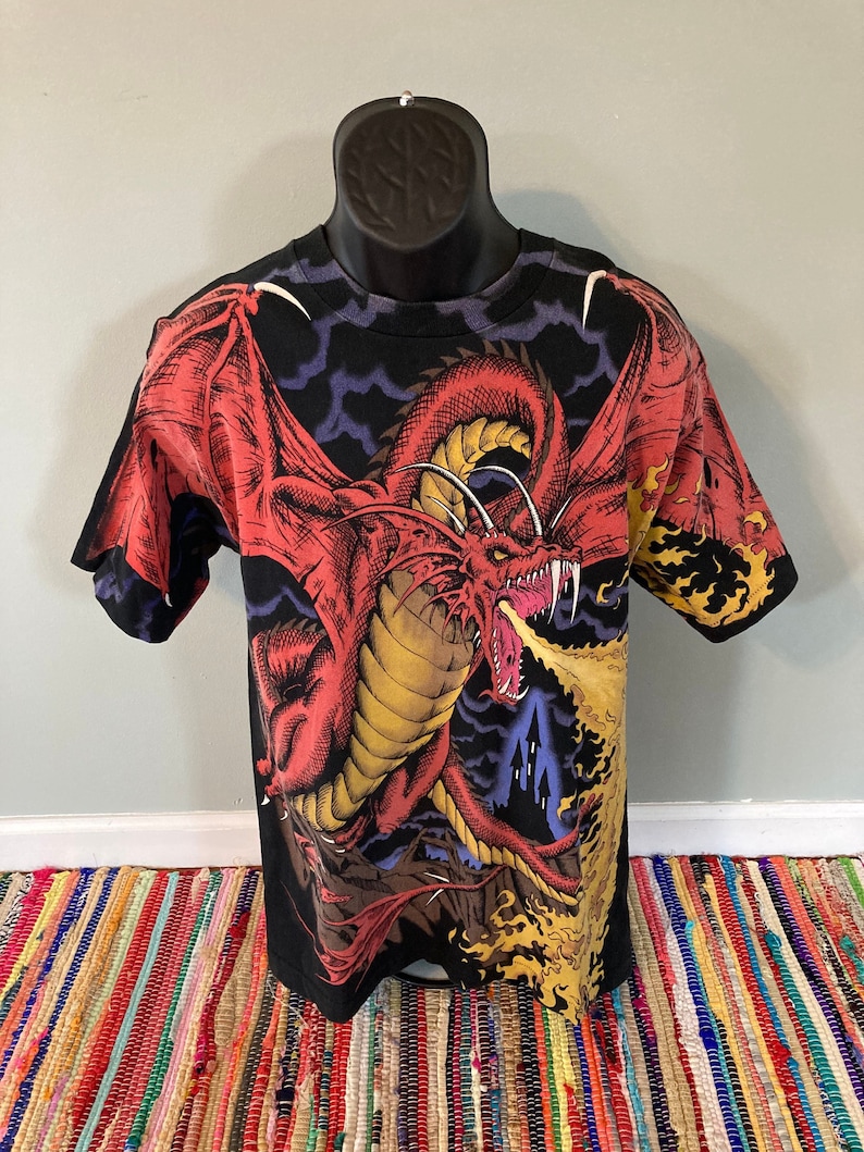 May include: Black t-shirt with a red dragon breathing fire in front of a dark castle. The dragon has yellow accents and is set against a dark blue and purple sky.