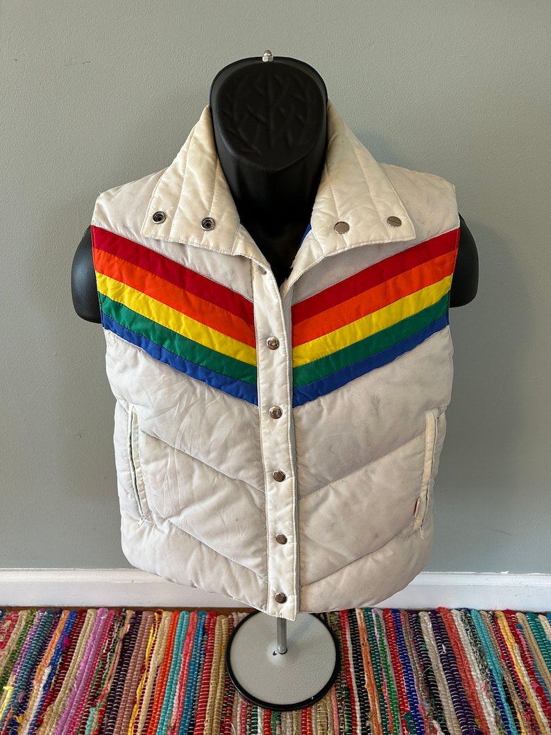 May include: A white puffer vest with a rainbow stripe design. The vest has a snap closure and a stand-up collar.