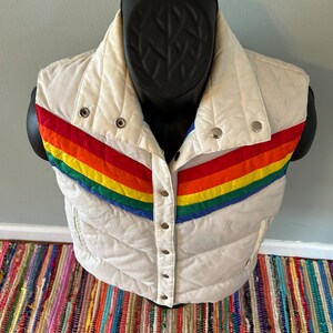 May include: White puffer vest with a rainbow stripe design across the front. The vest has a snap closure and is quilted.
