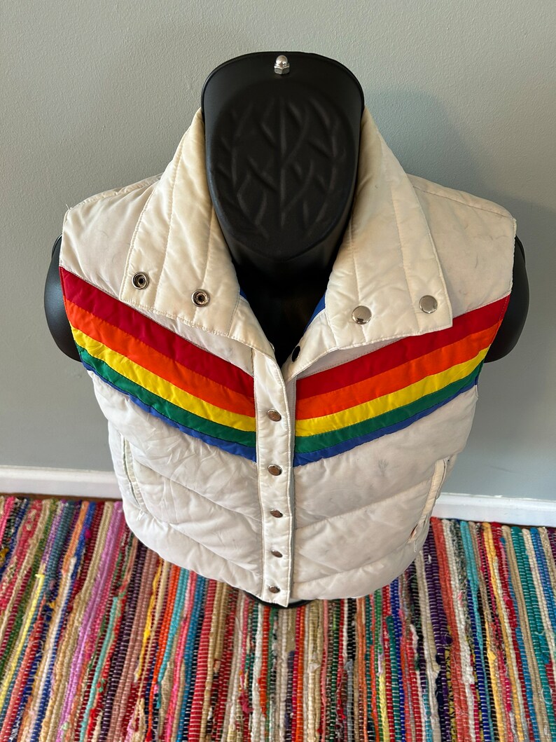 May include: White puffer vest with a rainbow stripe design across the front. The vest has a snap closure and is quilted.