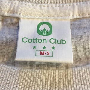 Pu&ograve; includere: Etichetta di una t-shirt di cotone bianco con un logo di pianta di cotone verde e il testo "Cotton Club" con tre stelle. L'etichetta riporta anche la taglia "M/5".