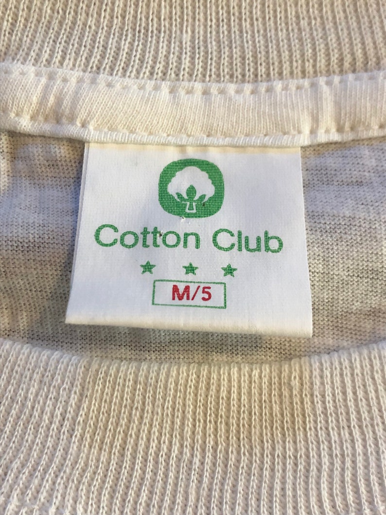 Pu&ograve; includere: Etichetta di una t-shirt di cotone bianco con un logo di pianta di cotone verde e il testo "Cotton Club" con tre stelle. L'etichetta riporta anche la taglia "M/5".