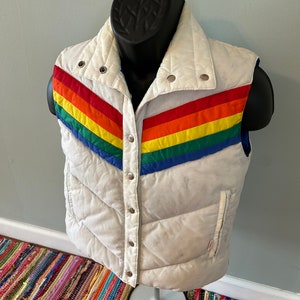 May include: White puffer vest with a rainbow stripe design on the front. The vest has a zipper closure and snap buttons.