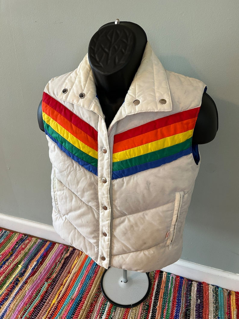 May include: White puffer vest with a rainbow stripe design on the front. The vest has a zipper closure and snap buttons.