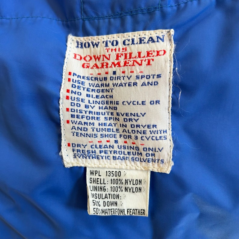 May include: A white care label with blue text detailing how to clean a down-filled garment. The label includes instructions for pre-scrubbing dirty spots, using warm water and detergent, and avoiding bleach. It also advises against using a lingerie cycle or spin dry, and recommends distributing the down evenly before washing. The label also includes instructions for dry cleaning using only fresh petroleum or synthetic base solvents. The label also includes the following information: WPL 13500, Shell: 100% Nylon, Lining: 100% Nylon, Insulation: 5% Down, 50% Waterfowl Feather.