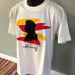 Pu&ograve; includere: Maglietta bianca con una silhouette nera di una testa e spalle di una persona. La silhouette &egrave; circondata da pennellate rosse, gialle e arancioni. Il testo "1791-1991" e "Mozart" sono stampati sotto la silhouette.