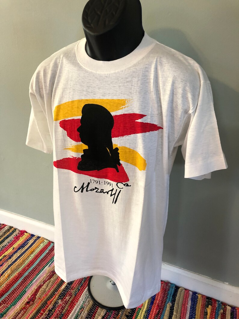 Pu&ograve; includere: Maglietta bianca con una silhouette nera di una testa e spalle di una persona. La silhouette &egrave; circondata da pennellate rosse, gialle e arancioni. Il testo "1791-1991" e "Mozart" sono stampati sotto la silhouette.