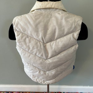 May include: A white puffer vest with a quilted design and a collared neckline. The vest is sleeveless and has a zipper closure.