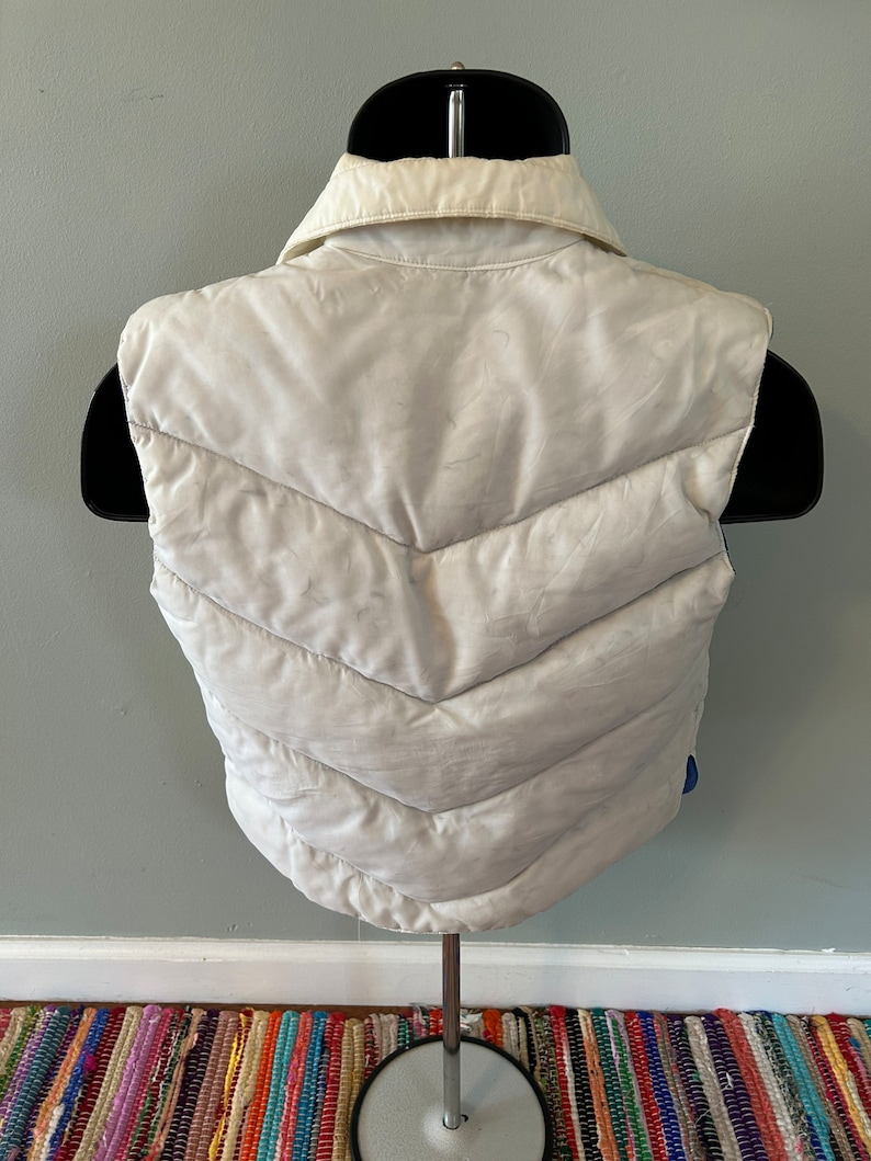 May include: A white puffer vest with a quilted design and a collared neckline. The vest is sleeveless and has a zipper closure.