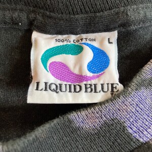 May include: A white tag with the text "100% Cotton" and "Liquid Blue" in black lettering. The tag has a blue, purple, and green swirl design. The tag is attached to a black shirt with a size "L" label.