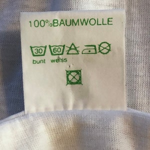 Pu&ograve; includere: Etichetta di abbigliamento bianca con testo e simboli verdi. L'etichetta riporta "100% Baumwolle" e include istruzioni di lavaggio per tessuti colorati e bianchi. I simboli indicano una temperatura massima di lavaggio di 30 gradi Celsius per tessuti colorati e 60 gradi Celsius per tessuti bianchi. L'etichetta include anche simboli per stirare e asciugare in asciugatrice.