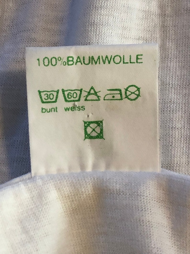 Pu&ograve; includere: Etichetta di abbigliamento bianca con testo e simboli verdi. L'etichetta riporta "100% Baumwolle" e include istruzioni di lavaggio per tessuti colorati e bianchi. I simboli indicano una temperatura massima di lavaggio di 30 gradi Celsius per tessuti colorati e 60 gradi Celsius per tessuti bianchi. L'etichetta include anche simboli per stirare e asciugare in asciugatrice.