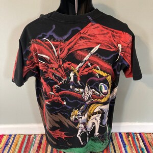 May include: Black t-shirt with a graphic print of a red dragon attacking a knight on horseback. The dragon has sharp claws and teeth, and the knight is holding a sword. The background is a dark stormy sky.
