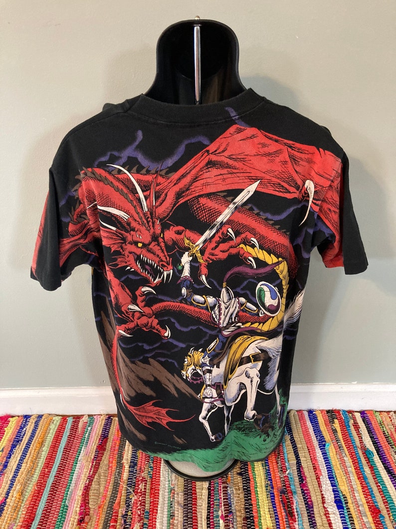 May include: Black t-shirt with a graphic print of a red dragon attacking a knight on horseback. The dragon has sharp claws and teeth, and the knight is holding a sword. The background is a dark stormy sky.