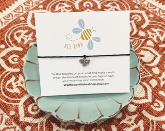 May include: A black string bracelet with a silver bee charm. The card reads "BEE KIND" and includes instructions to tie the bracelet on your wrist and make a wish. When the bracelet breaks in two, legend says your wish may soon come true. The card also includes the website WallflowerWishesShop.etsy.com.