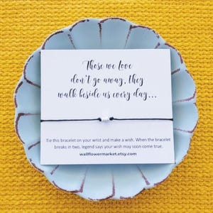 May include: A white card with black text that reads "Those we love don't go away, they walk beside us every day..."  The card is attached to a black string bracelet with a white bead. The bracelet is on a light blue plate with a scalloped edge. The plate is on a yellow surface.