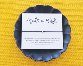 May include: A white card with black text that reads "Make a Wish".  A black string bracelet with silver beads is tied around the card. The card says "Tie this bracelet on your wrist and make a wish. When the bracelet breaks in two, legend says your wish may soon come true."