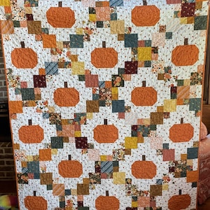 May include: A patchwork quilt featuring a repeating pattern of orange pumpkins with brown stems, set against a white background with small floral motifs. The design incorporates various square fabric blocks in shades of mustard, teal, and burgundy.