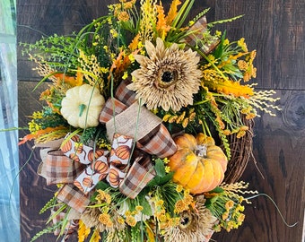 May include: A fall-themed wreath with a brown base, decorated with a variety of fall foliage, including yellow and orange leaves, small white flowers, and a large beige flower. The wreath also features a small white pumpkin and a larger orange pumpkin.