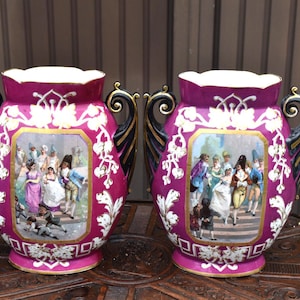 Pode incluir: Um par de vasos de cerâmica magenta com alças pretas e douradas em voluta. Cada vaso apresenta um painel retangular central retratando um grupo de pessoas com trajes históricos, rodeado por motivos florais brancos e detalhes dourados.