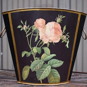 May include: A black metal wine cooler bucket with a gold-toned rim and base, featuring a floral illustration of pink roses and green leaves on the front. The bucket has two circular gold-toned side handles and is positioned next to a wine bottle.