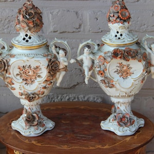 Könnte beinhalten: Paar antiker Porzellanvasen mit aufwendigen floralen und Putten-Details. Die Vasen sind weiß mit goldenen Akzenten und weisen erhabene Blumendesigns in Braun- und Orangetönen auf. Jede Vase hat eine dekorative Ober- und Unterseite.