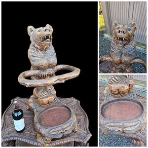 May include: A detailed, wooden bear-shaped umbrella stand. The bear is carved with an open mouth and sits atop a ring-shaped support. The stand has a lower tray and is intricately carved with floral and leaf patterns. A bottle of wine is next to the stand.