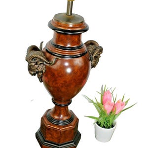 May include: A decorative table lamp with a brown, urn-shaped body and bronze ram's head handles. The lamp has a black base and trim, and a brass lamp holder. A small pot of pink tulips sits nearby.