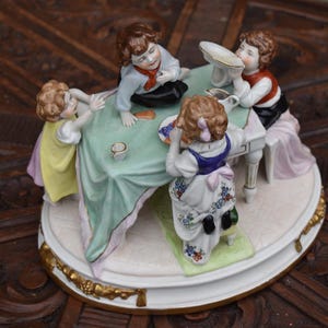 May include: A porcelain figurine depicting four children gathered around a table. The children are dressed in colorful clothing, with the table covered in a green cloth. The figurine is set on a decorative base with gold accents.