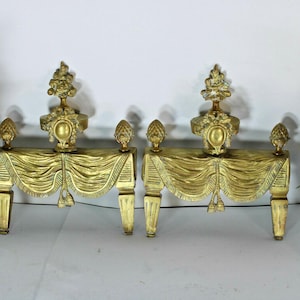 Puede incluir: Un par de morillos de chimenea de metal dorado con un diseño de tela drapeada y borlas, rematados con remates decorativos. Cada morillo tiene cuatro patas y un medallón central ovalado, con un acabado metálico de detalles intrincados.