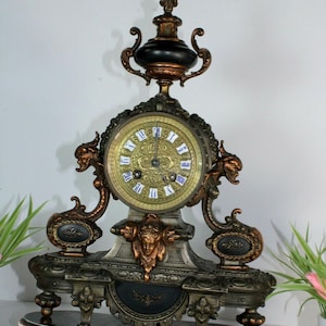 May include: A mantle clock featuring a circular gold-toned face with blue Roman numerals, set within a dark bronze-colored frame. The structure includes ornate scrollwork, a central face relief, and a black finial top, all resting on a wide base.