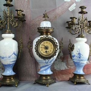 May include: A three-piece set featuring a central clock and two flanking candelabras. Each piece is crafted from white ceramic with blue landscape illustrations, accented by ornate brass bases, handles, and candle holders.