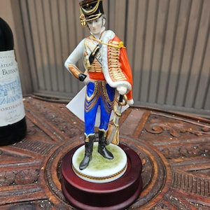 Puede incluir: Figura de porcelana de un soldado con uniforme militar. La figura está sobre una base de madera redonda. El soldado viste una chaqueta blanca, pantalones azules y un abrigo rojo. El sombrero es negro con detalles dorados.