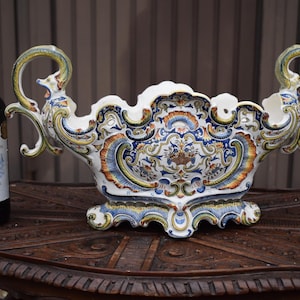 Pode incluir: Um grande centro de mesa em cerâmica branca com intrincados motivos florais e volutas pintados em azul, amarelo e laranja. A taça possui duas pegas curvas e ornamentadas em forma de cabeças de animais estilizadas e um rebordo festonado.