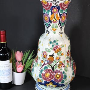 Może przedstawiać: Wysoki, pękaty wazon ceramiczny z falistym brzegiem, ozdobiony ręcznie malowanymi wzorami kwiatowymi w kolorach czerwonym, pomarańczowym, żółtym i zielonym oraz wizerunkami małych ptaków siedzących na gałęziach na białym tle na okrągłej podstawie.